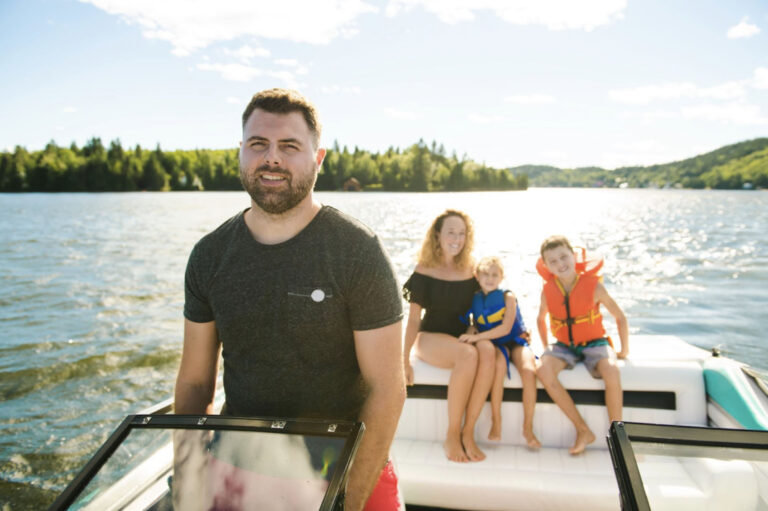 Man driving a boat with a smiling family in the background, including two children wearing life jackets, enjoying a sunny day on the water.